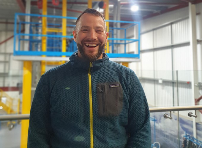 A man in a blue fleece jacket stands indoors in front of industrial equipment, smiling broadly at the camera.