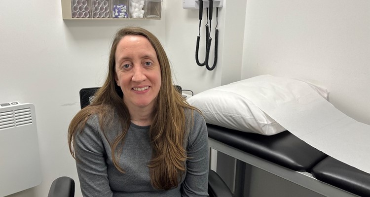 A woman with long hair sits in a medical examination room with a bed and medical equipment in the background. She is wearing a gray shirt and smiling at the camera.