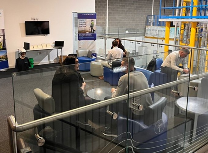 A group of people are seated and conversing in a modern, open-plan office space, featuring glass dividers, banners, and a wall-mounted TV.