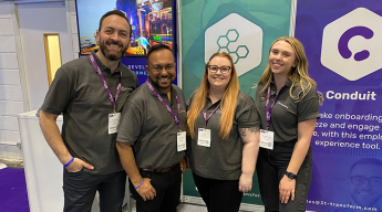 Four people wearing matching shirts stand together, smiling, in front of a display booth featuring promotional banners at an event or conference.