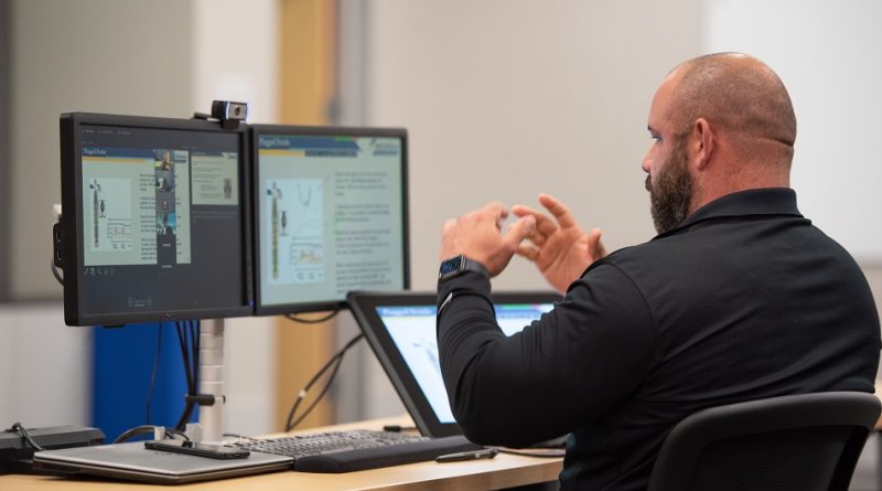 A person sitting at a desk with dual monitors and a tablet, gesturing with their hands, possibly during an online meeting or presentation.