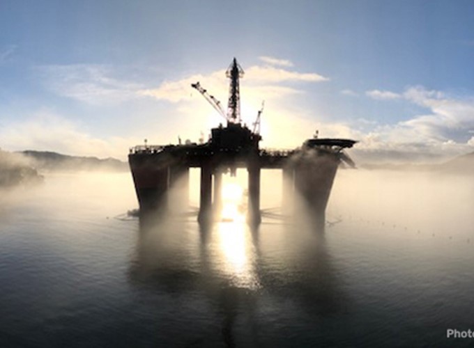 An offshore oil platform stands in the middle of the water with the sun setting behind it, casting a silhouette and reflecting on the water's surface. Mist surrounds the platform.