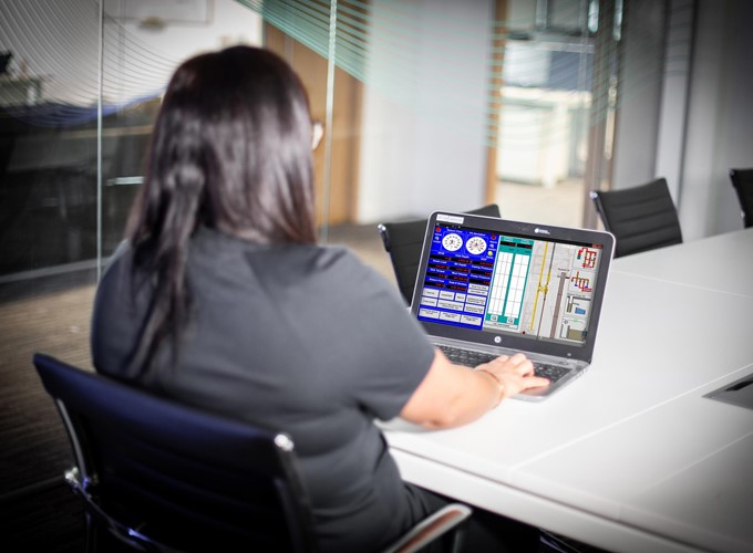A person with long dark hair sits at a conference table, working on a laptop with a technical interface displayed on the screen.