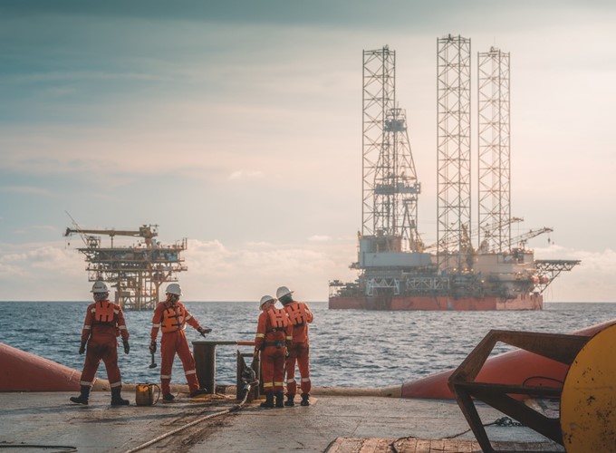 Four workers in orange suits stand on an offshore platform with oil rigs visible in the background under a cloudy sky.