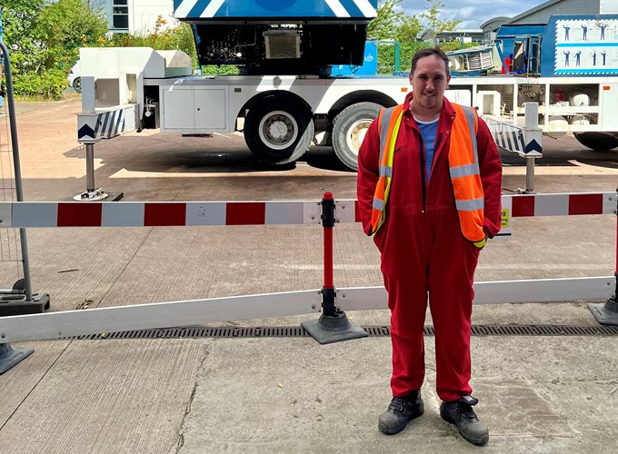 Person in a red jumpsuit and reflective safety vest stands in front of a barrier, with a large industrial vehicle in the background.