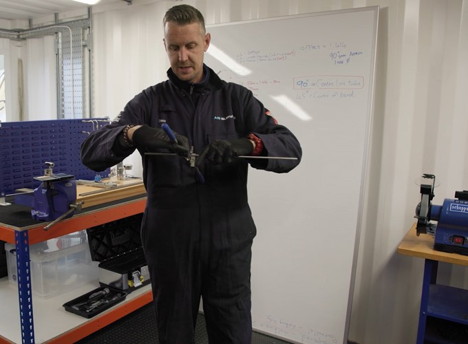 A person in a workshop is using a hand tool to work on a piece of metal, with a whiteboard in the background displaying some notes. Shelving and various tools are visible around the workshop.