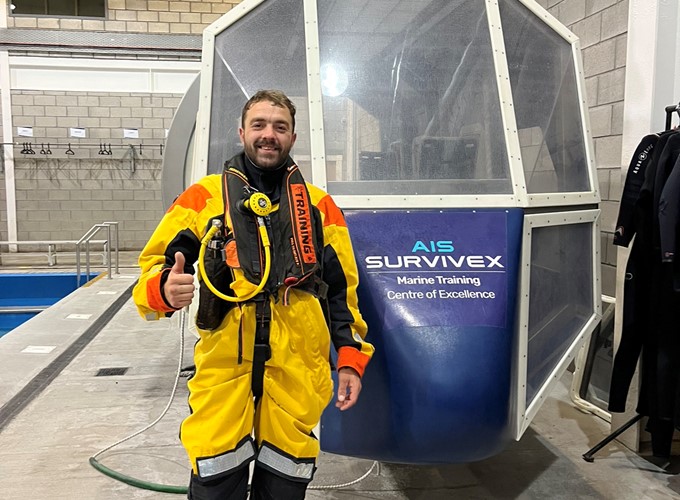 A man in a yellow and orange survival suit stands indoors, giving a thumbs-up. He is in front of a training simulator labeled 