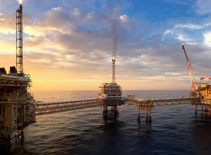 An offshore oil platform at sunset with a network of connected walkways over the ocean and a crane on the right side.