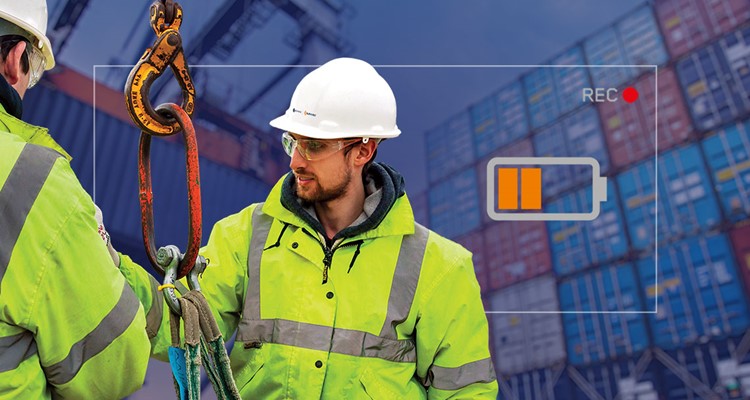 Two workers in high-visibility jackets and hard hats operate lifting equipment at a container yard, with an on-screen battery icon and a red recording indicator overlay.