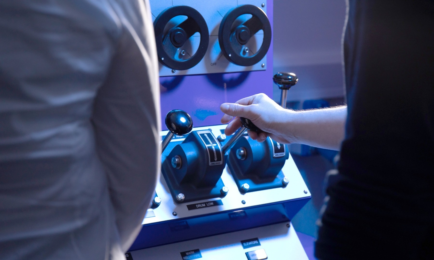 Two individuals operate a control panel with various levers and dials. One person's hand is shown gripping a lever.