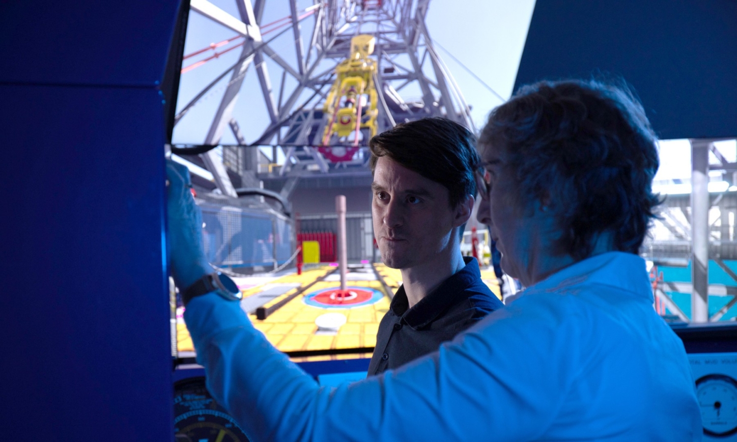 Two men in a control room operate a simulator with a large screen displaying the interior of an industrial setting. One is pointing while the other observes intently.