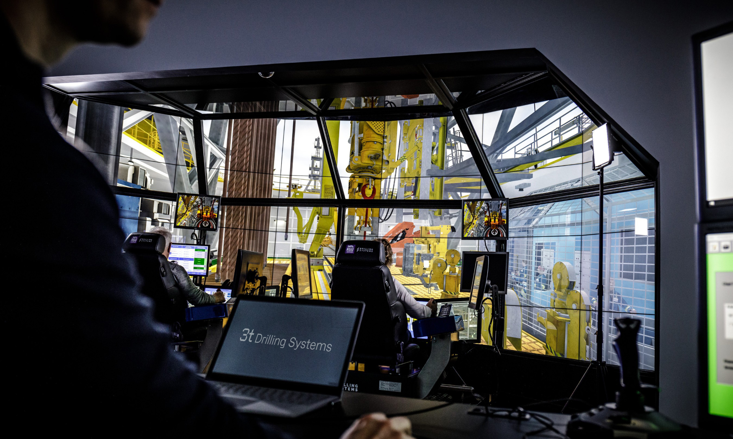 People operating a drilling simulator with multiple screens displaying various control panels and machinery. A person in the foreground is working on a laptop that says 