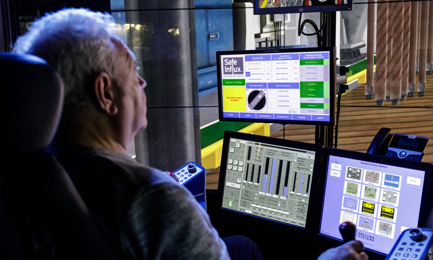 A man operates a control board with multiple screens and buttons. The screens display various charts, controls, and information, including one labeled 