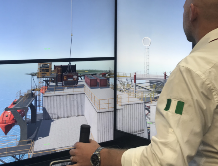 A man operates a control joystick while viewing a detailed 3D rendering of an offshore platform on a large screen.