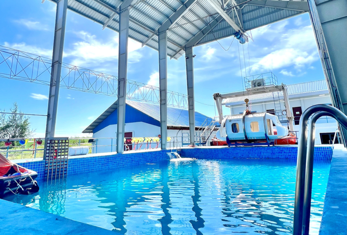 An indoor pool with a metallic roof structure overhead. A large piece of equipment is positioned beside the pool, and a blue-roofed building is visible in the background.