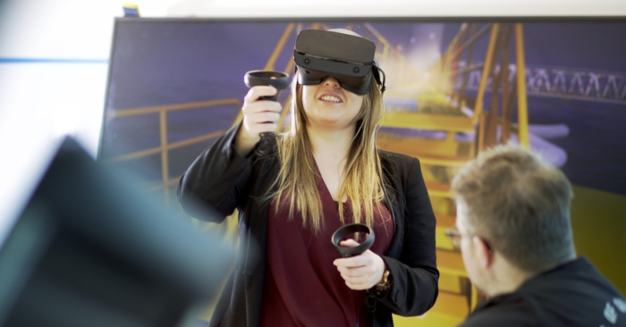A woman wearing a virtual reality headset and holding controllers interacts with a digital simulation, while a man observes in the foreground.