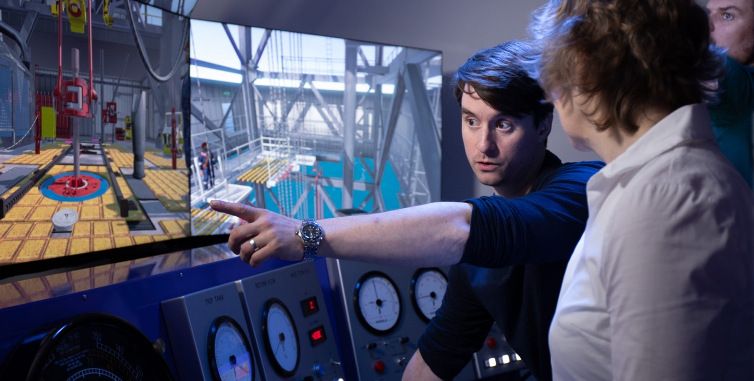 A person points at a screen displaying an industrial setting while explaining something to another person. Dials and gauges are visible on a control panel in the foreground.
