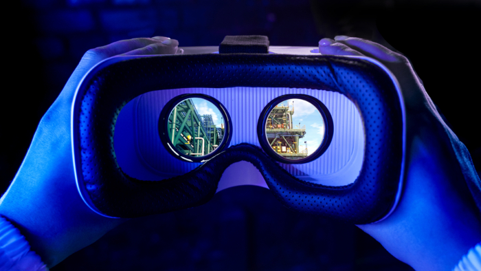 Hands holding a virtual reality headset with a view of industrial structures seen through the lenses.