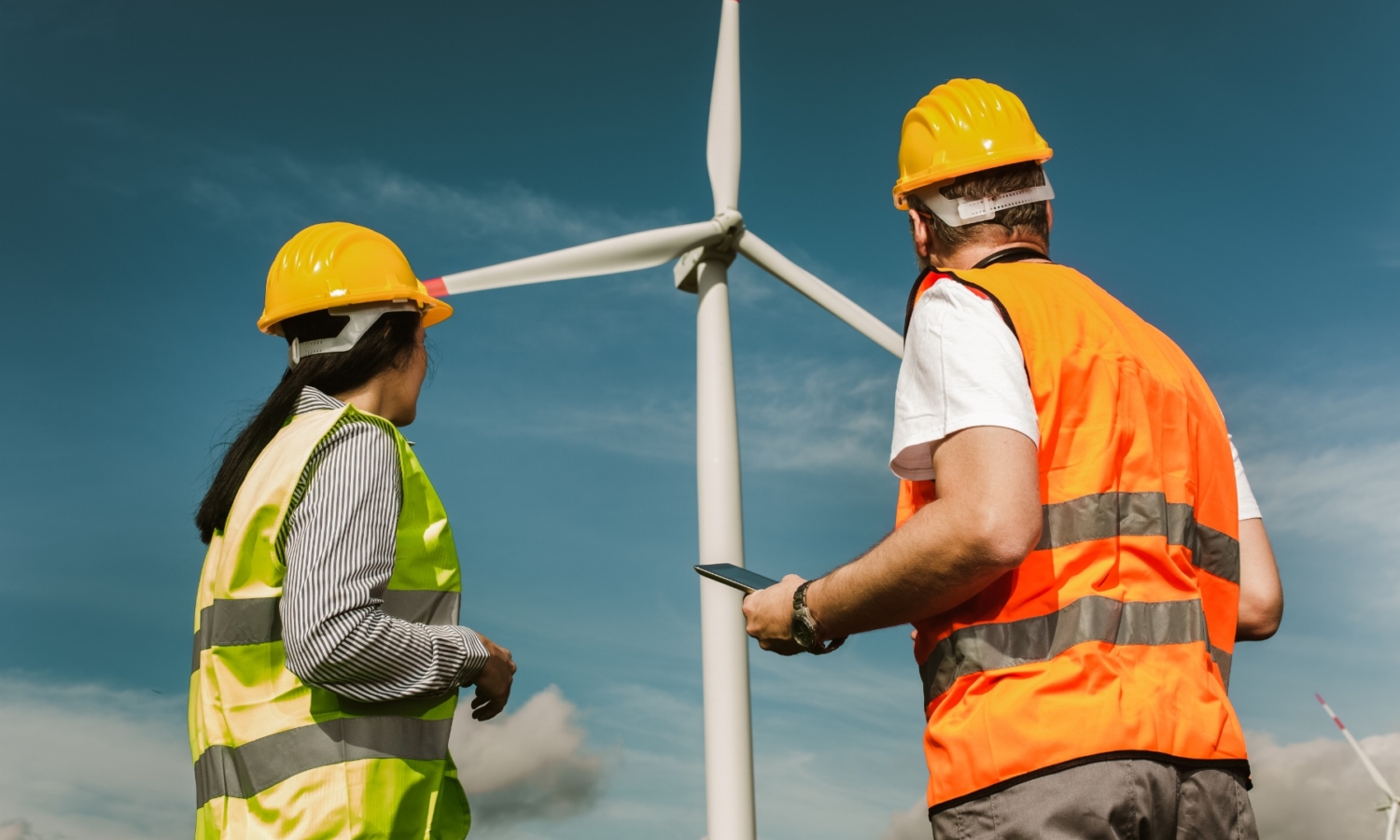 Two workers wearing hard hats and reflective vests stand near a large wind turbine, observing and possibly inspecting it under a clear blue sky.