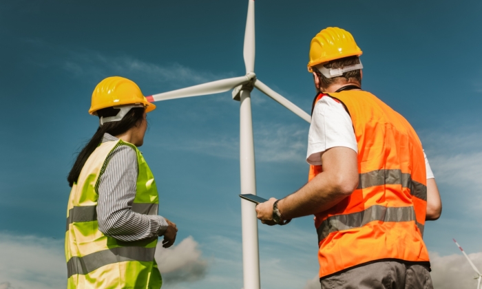 Two workers wearing hard hats and reflective vests stand near a large wind turbine, observing and possibly inspecting it under a clear blue sky.