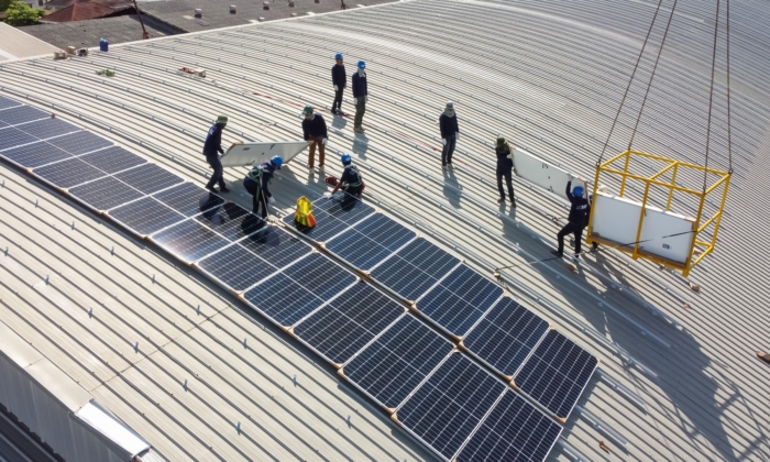 Workers installing solar panels on a large, ribbed metal rooftop, using a crane to lift equipment.