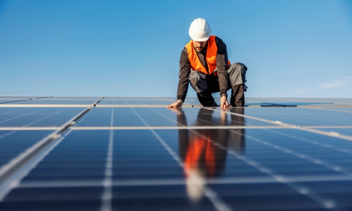 Worker in a safety vest and helmet installs solar panels on a sunny day.