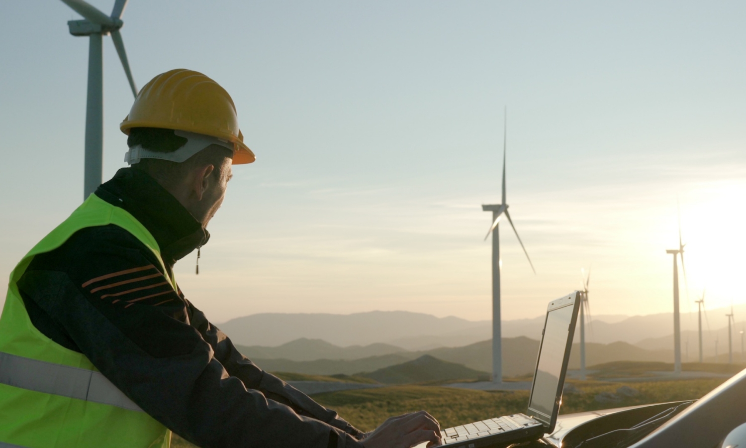 Person in a hard hat and safety vest using a laptop outdoors near wind turbines, with a scenic hilly landscape in the background during sunset.