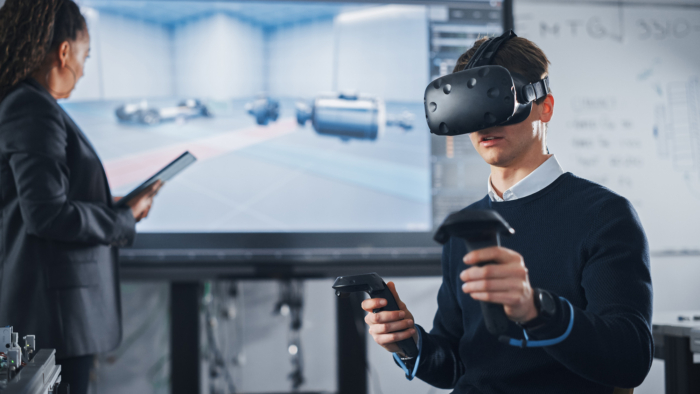 A man wearing a virtual reality headset and holding controllers sits in front of a large screen displaying a virtual environment. A woman stands nearby holding a tablet, viewing the large screen.