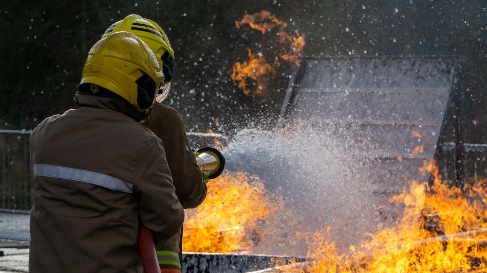 Two firefighters in protective gear aim a hose to extinguish a fire, with flames and smoke billowing around.