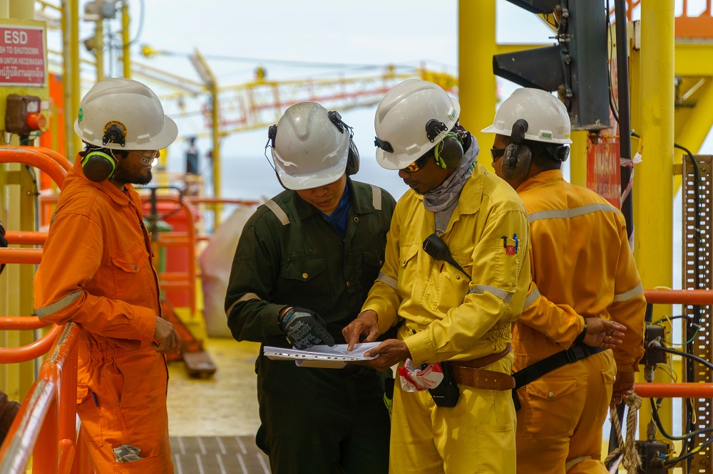 Four workers in safety gear review a document on an offshore oil rig. Two in orange coveralls, one in black, and one in yellow, discuss the contents while standing next to orange railings.
