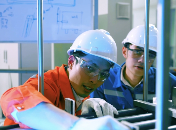 Two workers in hard hats and safety glasses are examining machinery in a facility. One worker in an orange jumpsuit is pointing, while the other in a blue jumpsuit observes. A whiteboard is visible behind them.