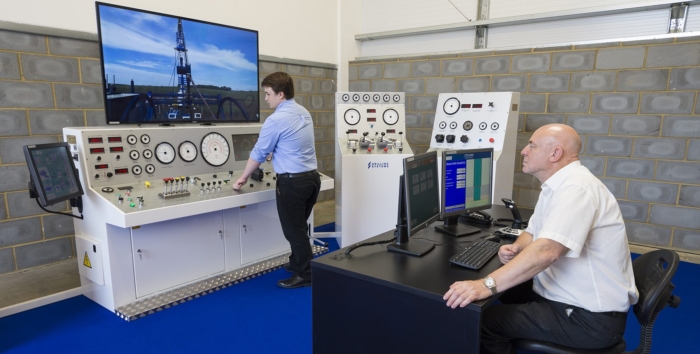 Two men operate an oil and gas drilling simulator in a control room, with one seated at a computer desk and the other standing at control panels. A large screen displays a drilling rig.