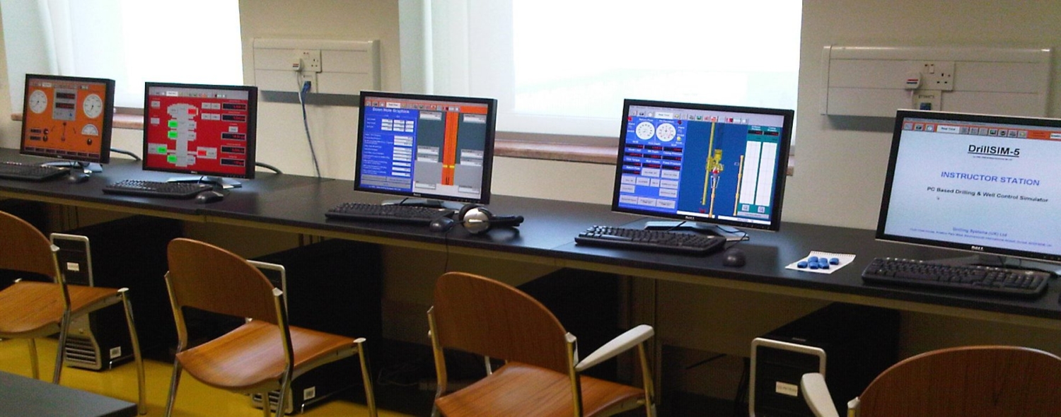 A row of five computer workstations with various software interfaces displayed on the monitors. There are keyboards, mice, and headsets at each station, with wooden chairs facing the monitors.
