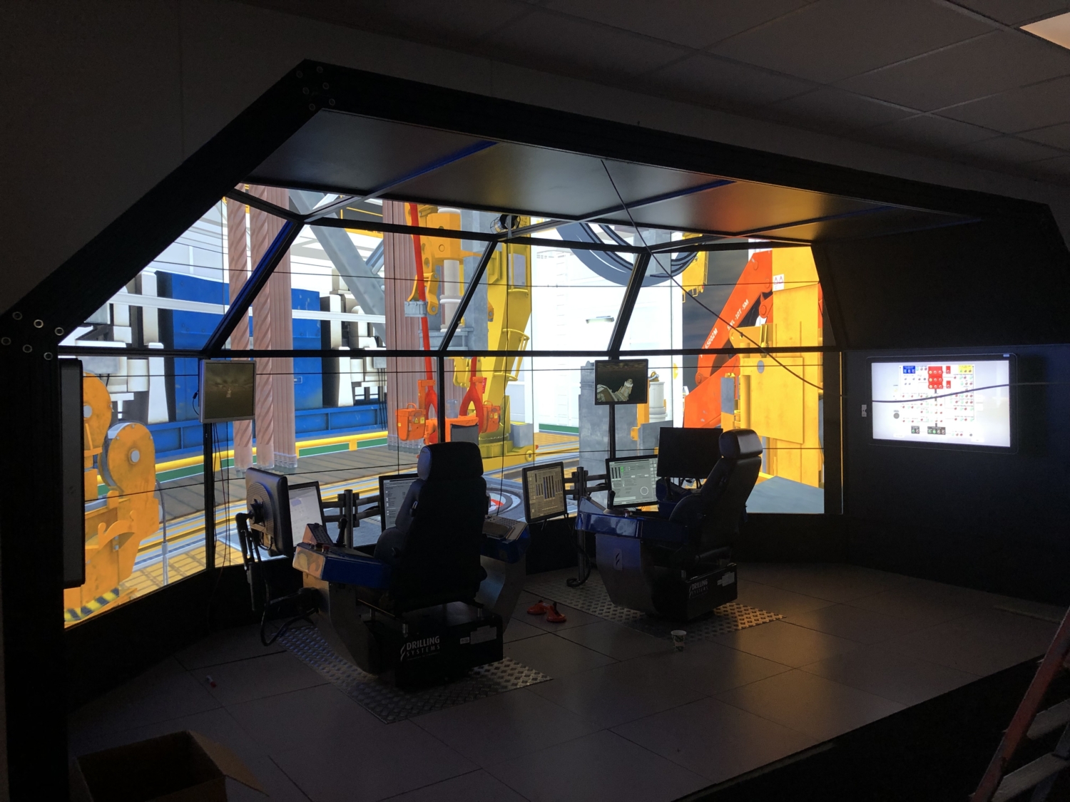 A modern simulation room with two control stations surrounded by large screen displays depicting an industrial environment.