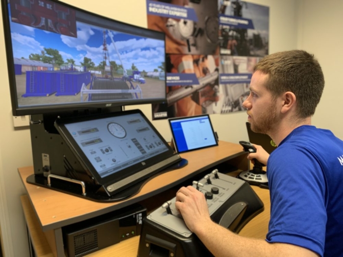 A person operates a control simulator with multiple screens displaying graphics and controls, simulating an industrial environment, in a training room with posters on the wall.