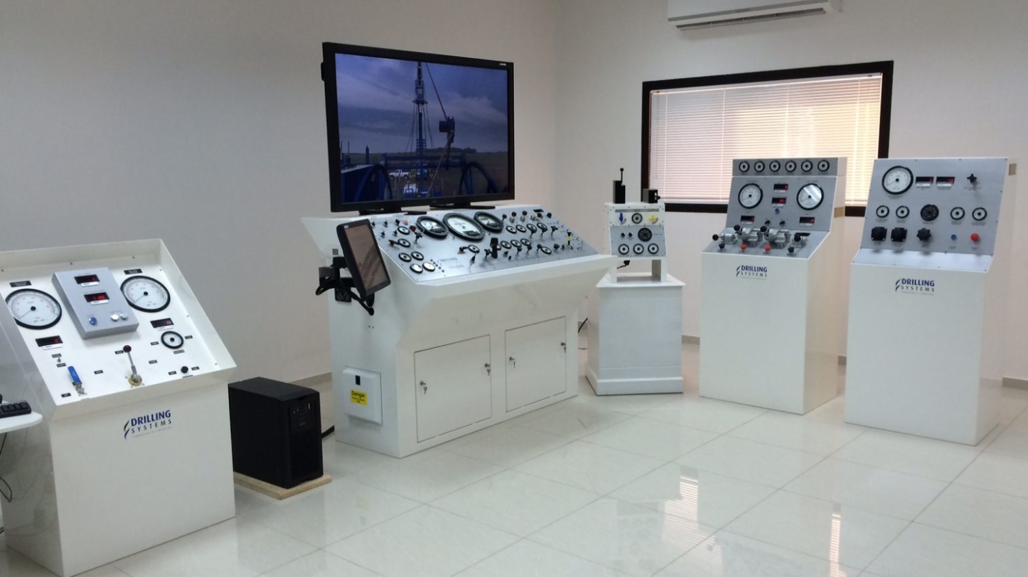 A room with multiple control panels, gauges, and a large screen on the wall. The equipment is arranged on white stands and cabinets, and the floor is tiled.