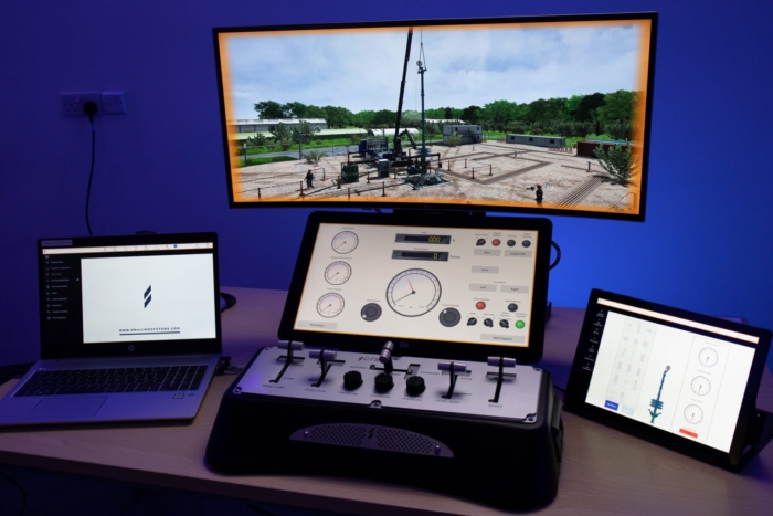 A workstation with two laptops, a central control panel, and a tablet displaying various data and interfaces. A large screen above shows an outdoor scene with machinery and structures.