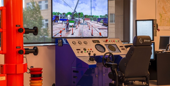 A training room with a control panel, large screen displaying an industrial scene, black chair, and various safety equipment on the left.