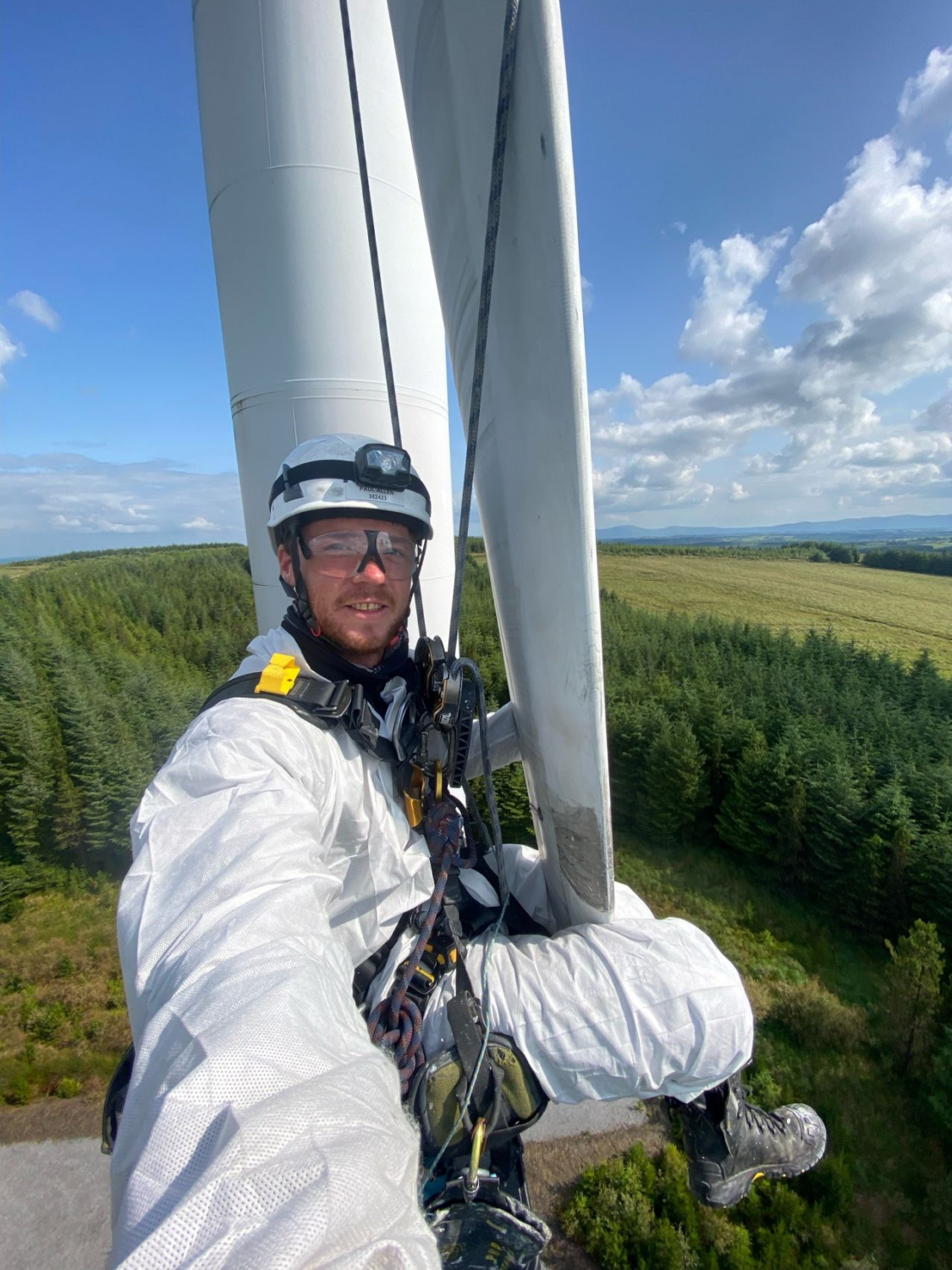 A person in protective gear and helmet is suspended by ropes on a wind turbine, with a forested landscape and clear sky in the background.