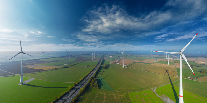 Aerial view of a wind farm with multiple wind turbines spread across a landscape of green fields and a road running through the middle under a partly cloudy sky, showcasing the transformation from military to wind energy initiatives.
