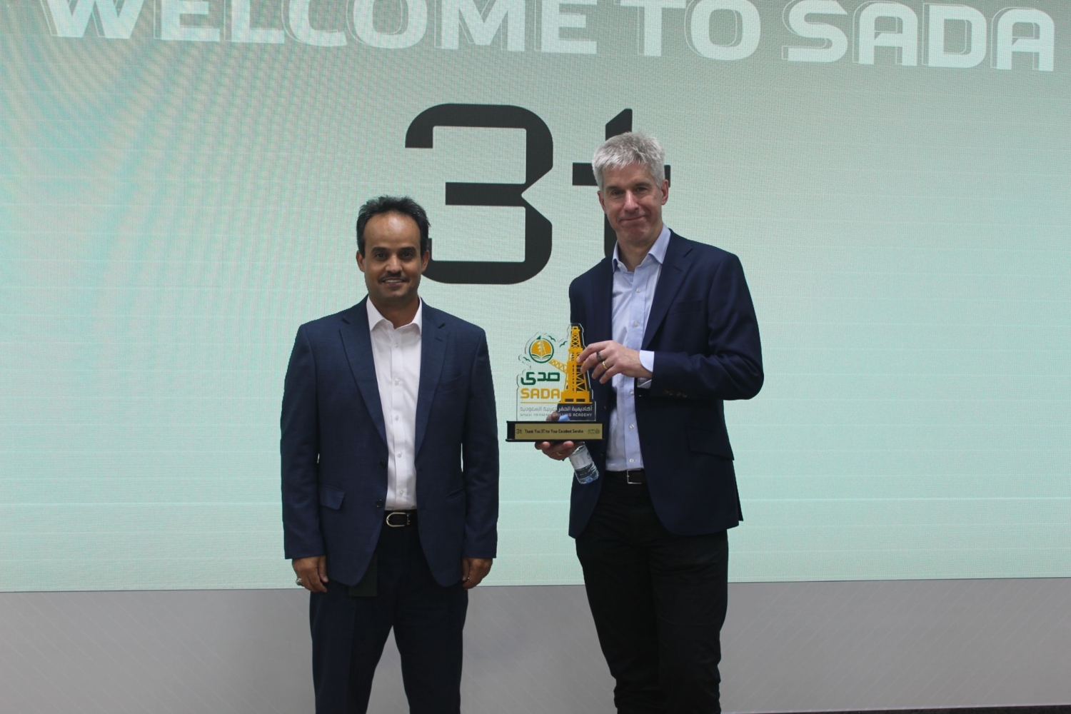 Two men in suits stand on a stage, one proudly holding an award. The screen behind them displays the text 