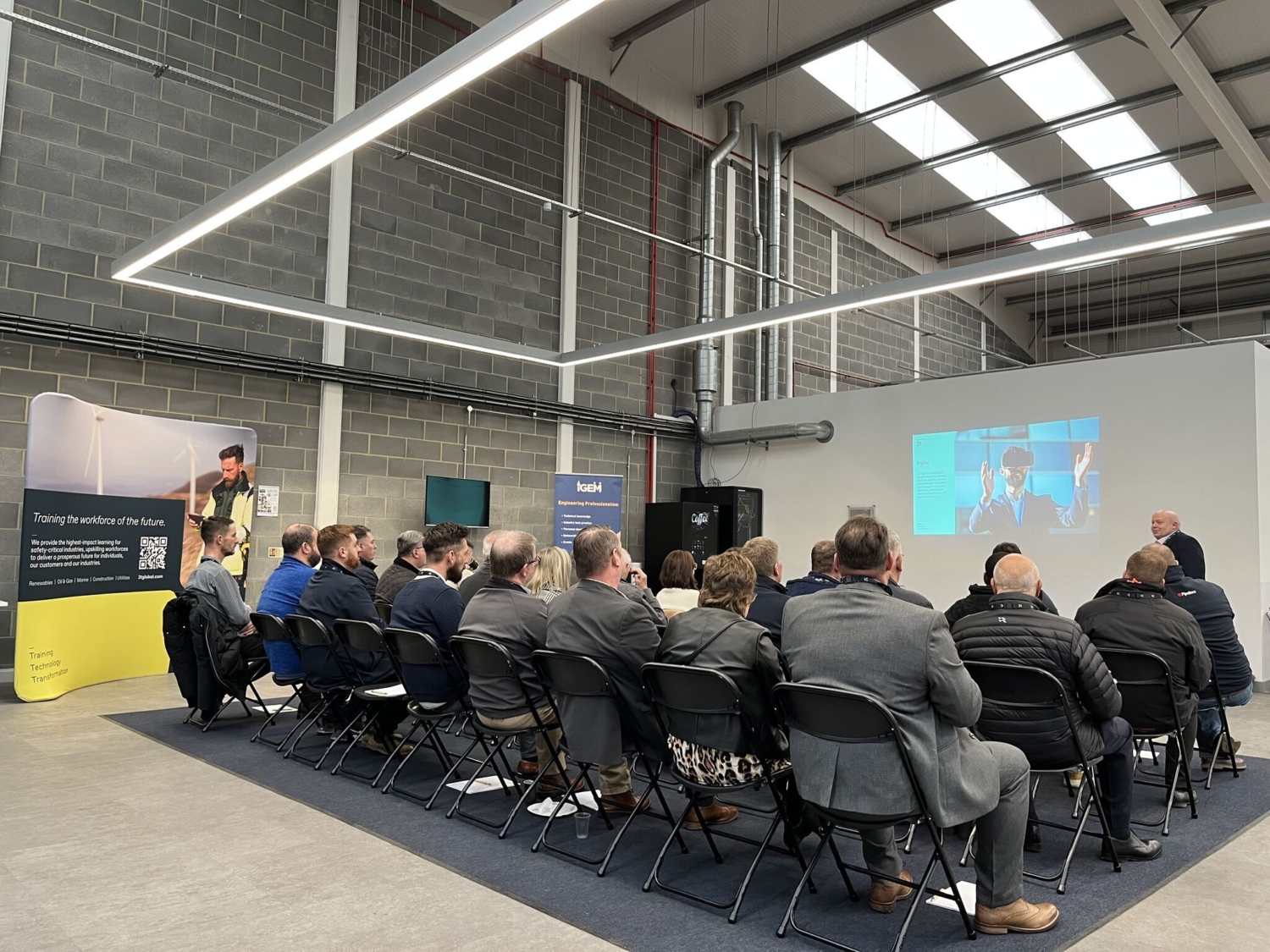 A group of people in rows attentively engages in a training session, facing a speaker and a large screen, all within the sleek ambiance of a modern industrial-looking conference room.