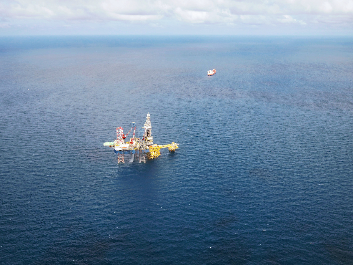 Aerial view of an offshore oil platform in the ocean, with a distant ship on the horizon under a partly cloudy sky, subtly illustrating nature's delicate balance amid the relentless training of industry.