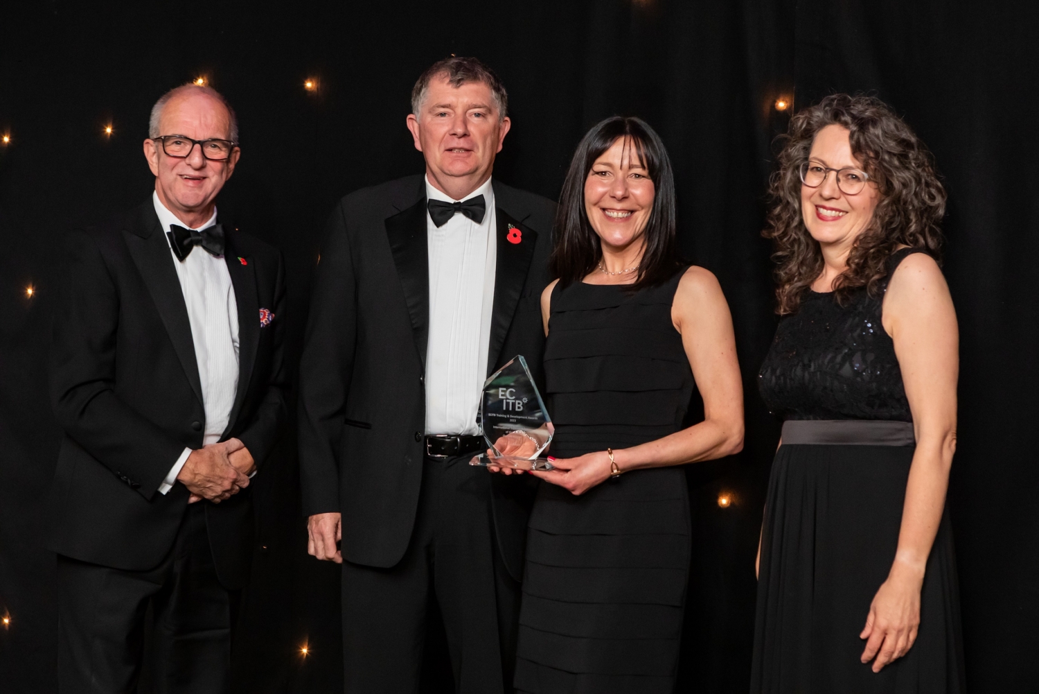 Four people in formal attire stand together against a black backdrop, showcasing their achievements after intensive training. The woman second from the right proudly holds a glass award as evidence of her success.