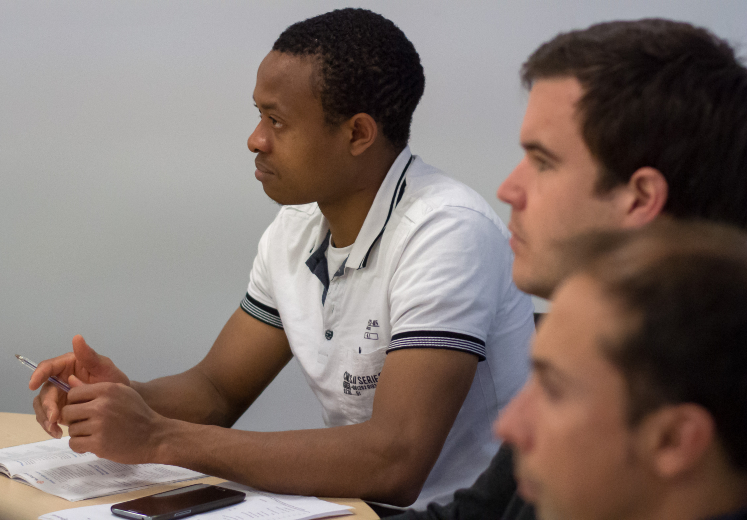 Three people are seated at a table, intently focusing on something out of view—perhaps a training session. One wears a white shirt while the others are partially visible. Papers and a phone lie scattered across the table.