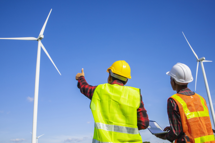 Two workers in safety gear stand outdoors, observing wind turbines against a clear blue sky. One points upward while the other holds a laptop, highlighting the importance of ongoing training in mastering these renewable energy systems.