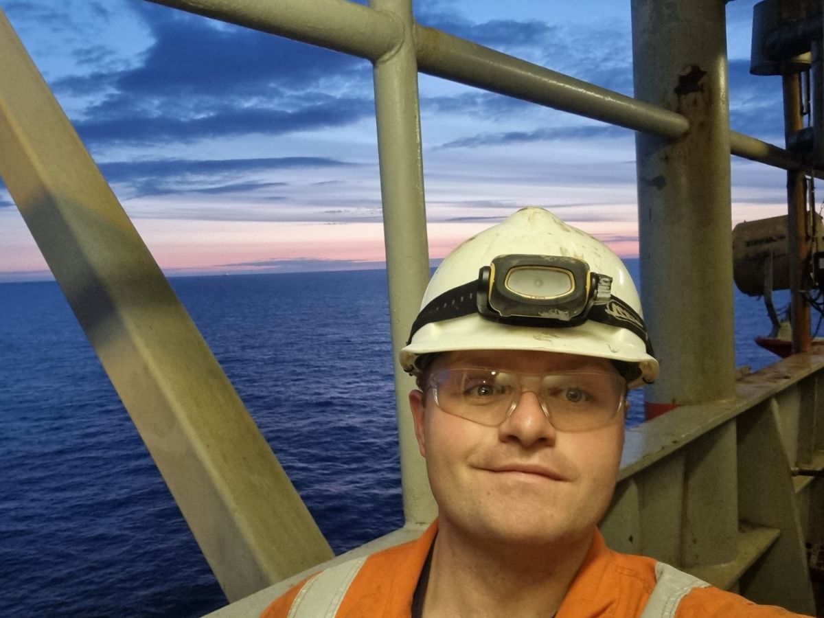 man in an orange jumpsuit, safety goggles and a white helmet smiling in front of a sunset offshore