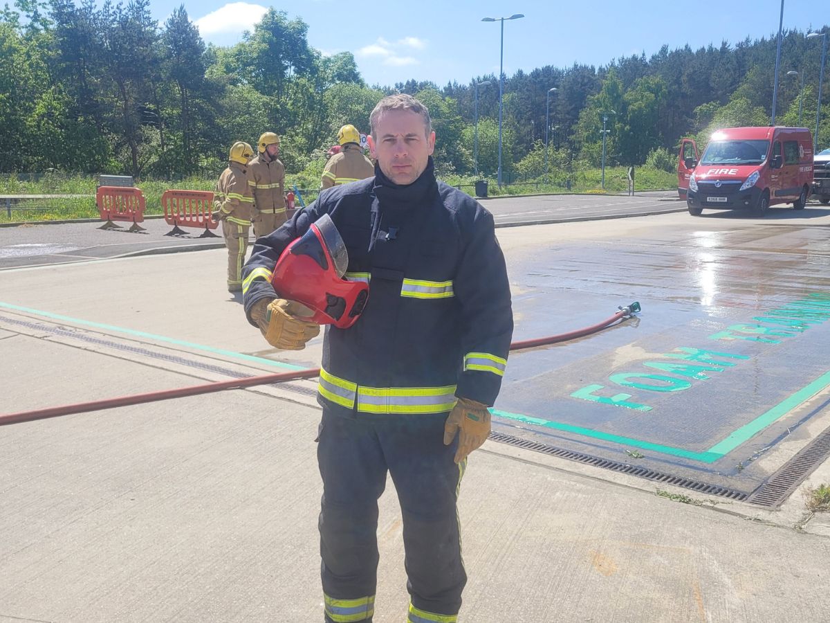 A firefighter holding a helmet. In the background are three more firefighters and a hose on the ground.