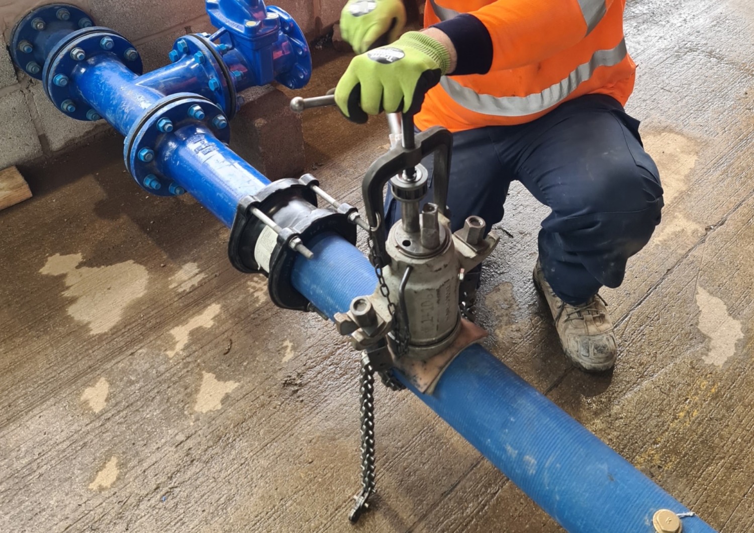 A worker in an orange high-visibility jacket and green gloves, undergoing construction training, operates a tool on a blue pipe with water valves indoors.