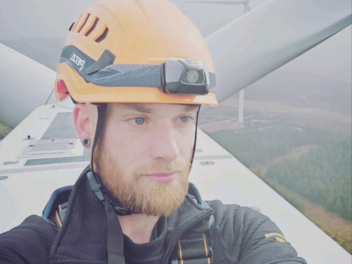 wind technician with orange helmet on the top of a wind turbine. In the background are the blades and countryside.